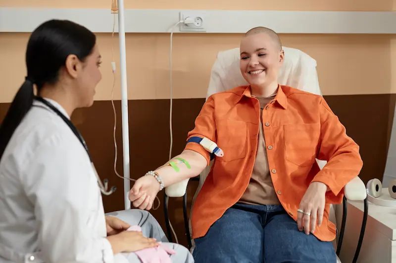 Женщина проходит курс химиотерапии: улыбающаяся пациентка с бритой головой получает инфузию и общается с врачом.